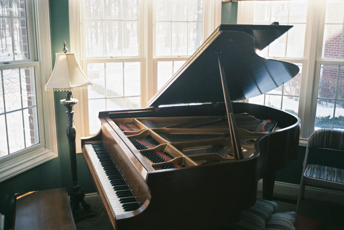 A mahogany grand piano with its lid raised in a bright bay window beside a standard lamp