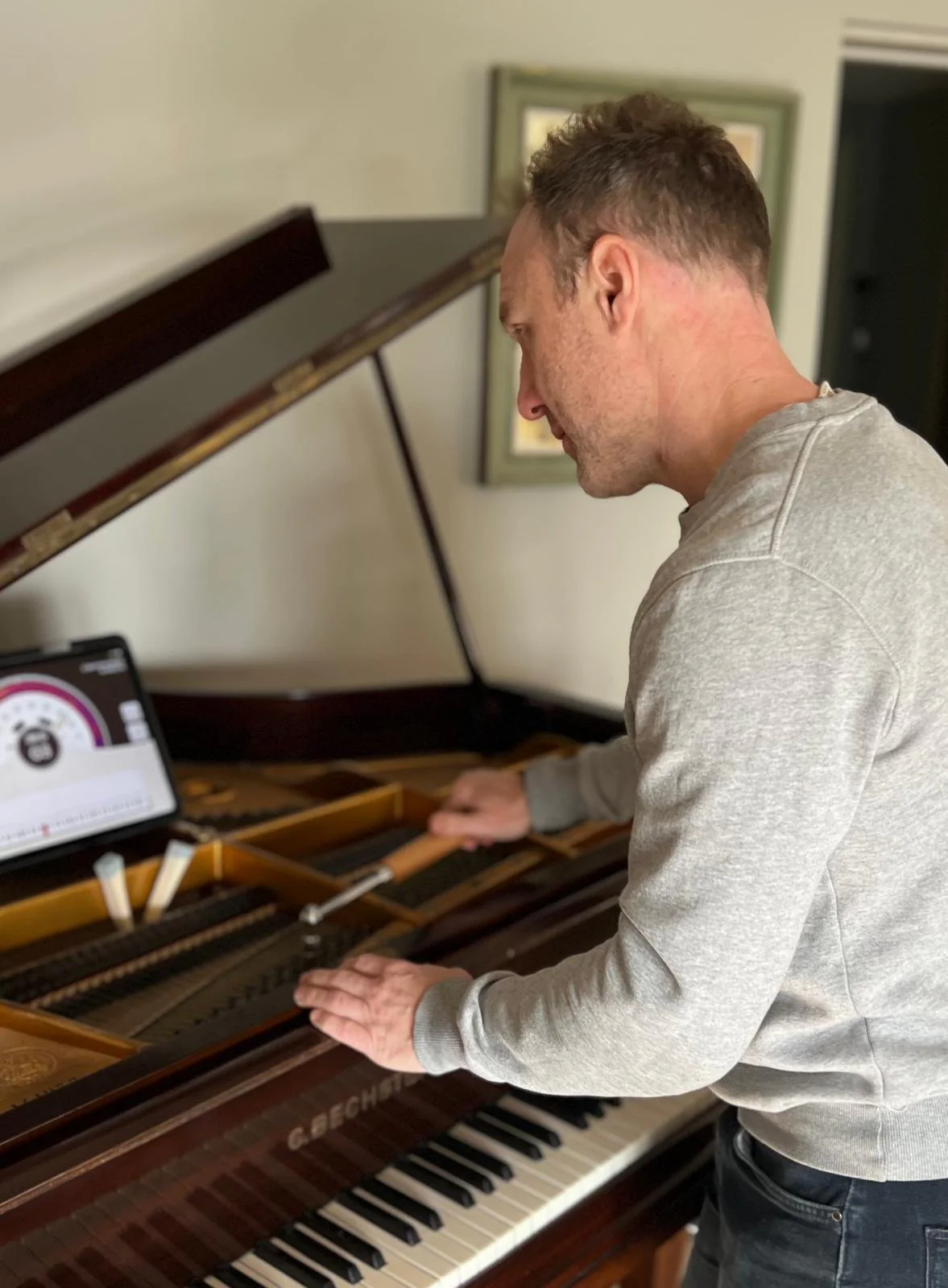 Pete Hills tuning a C. Bechstein grand piano in a client's home, tuning lever on the pins, felt mutes in the strings and a tuning app on a tablet