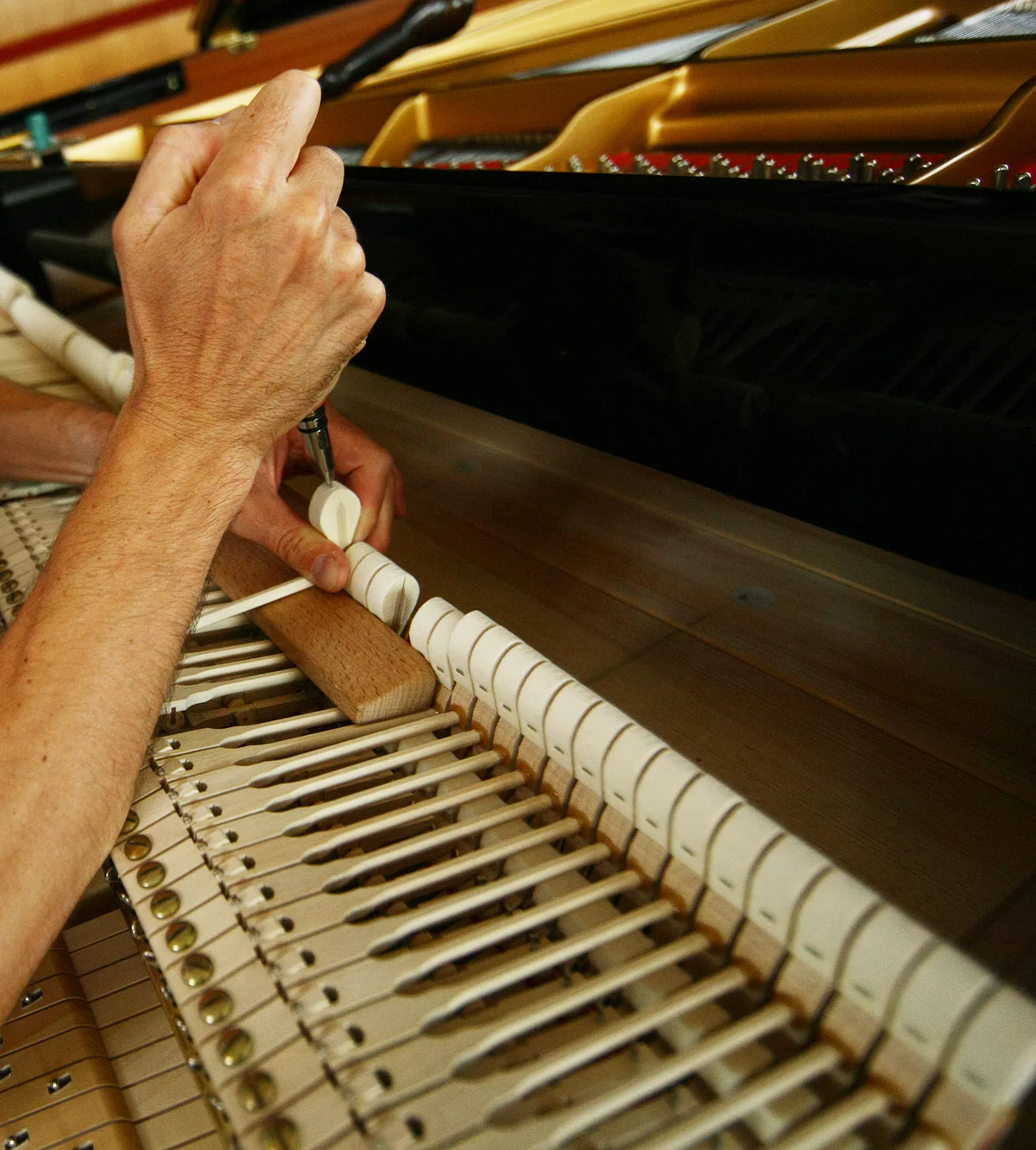A technician's hands voicing a felt hammer with a needle tool on a grand piano action pulled out onto the keybed