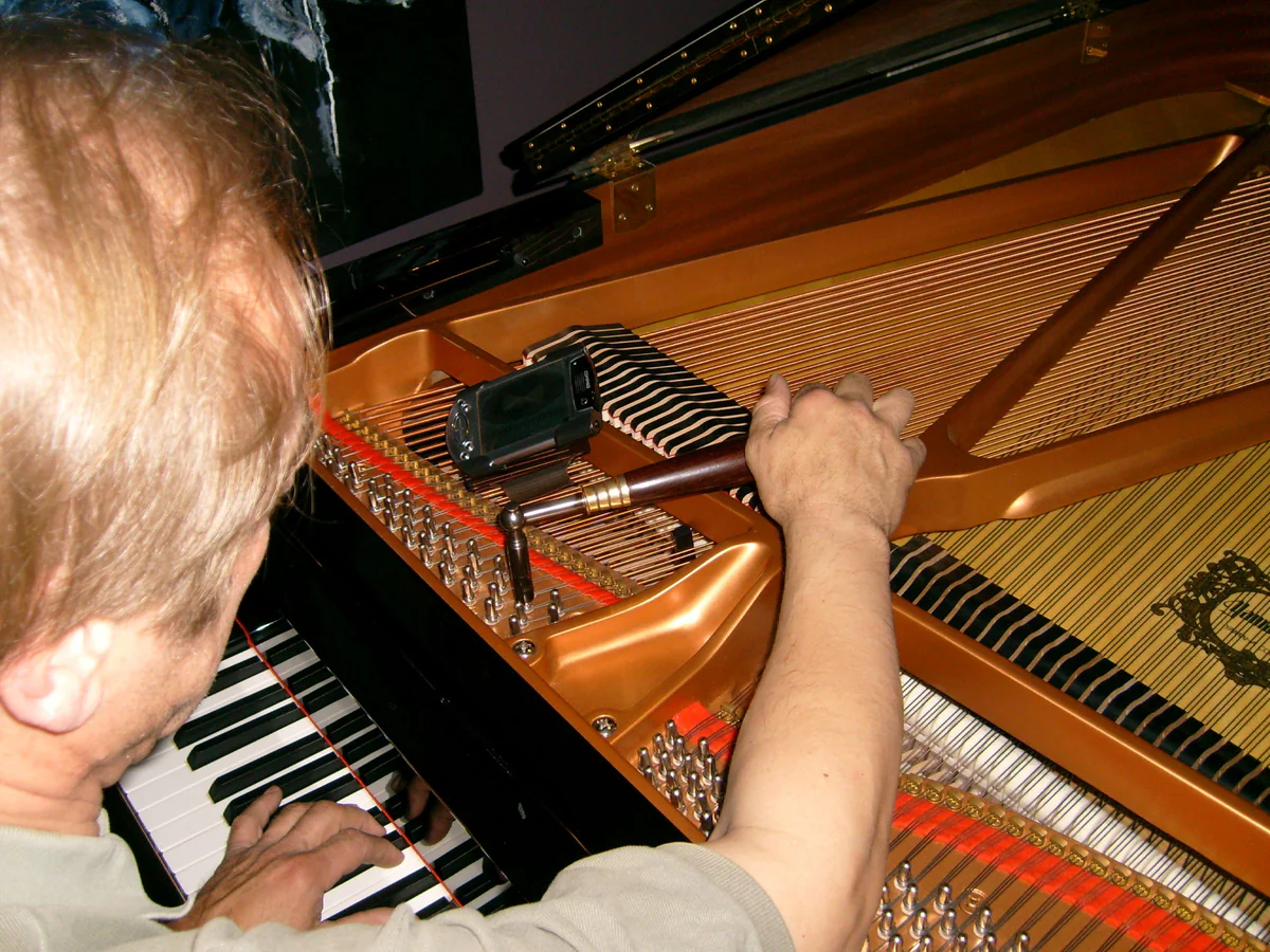 Over-the-shoulder view of a technician tuning a grand piano: tuning lever on the pins, felt mute strip in the strings and an electronic tuner resting on the iron frame